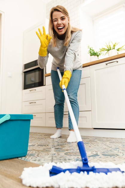 joyful young woman housewife in gloves gesturing ok sign and winking while mopping floor at kitchen