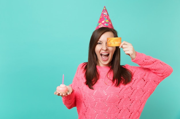 Joyful young woman in knitted pink sweater, birthday hat holding in hand cake with candle, covering eye with credit card isolated on blue wall background. People lifestyle concept. Mock up copy space.