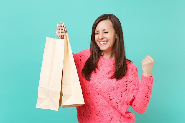 Joyful young woman in knitted pink sweater doing winner gesture, holding packages bags with purchases after shopping isolated on blue wall background. People lifestyle concept. Mock up copy space.