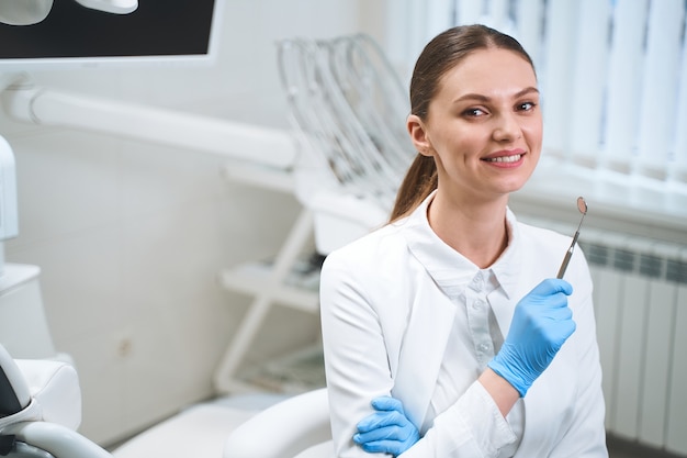 Foto gioiosa giovane donna in uniforme medica è in piedi nel suo ufficio moderno e tiene in mano uno specchio dentale.