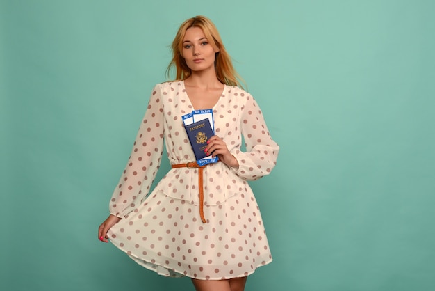 Joyful young woman in polka dot dress is holding airline tickets with a passport on a blue background.