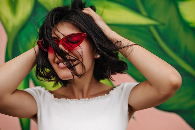 Joyful young woman posing near green graffiti Charming brunette girl in sunglasses touching hair