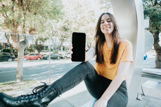 Joyful young woman showing display of mobile phone with screen to copy space Advertisement marketing and publicity Young adult smiling isolated in urban scenery in the city looking at camera