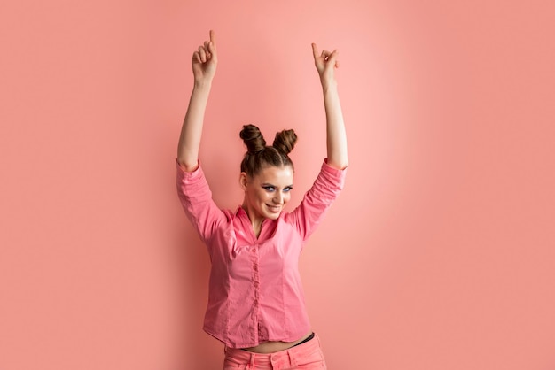 Joyful young woman standing with two bun hairstyle in pink skirt stands with arms raised up and points fingers