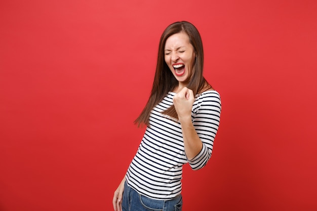 Joyful young woman with closed eyes in striped clothes clenching fist like winner and screaming 