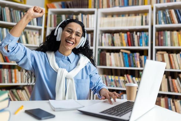 Photo joyful young woman with headphones cheering in front of a laptop experiencing triumph in a library