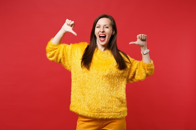 Joyful young woman in yellow fur sweater keeping mouth wide open, pointing thumbs on herself isolated on red wall background in studio. People sincere emotions, lifestyle concept. Mock up copy space.