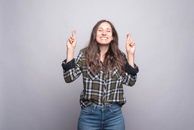 Joyfull young woman smiling and standing with crossed finger over grey wall