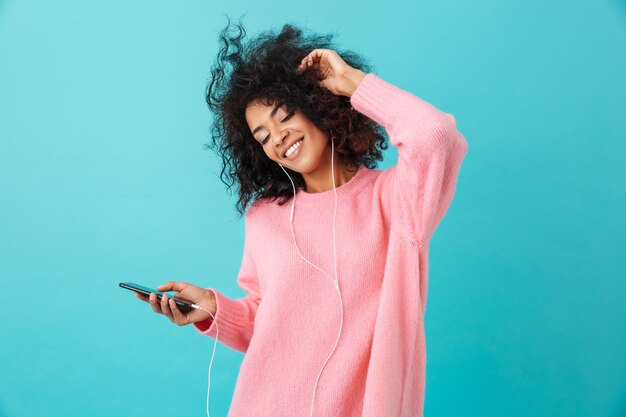 Joyous american woman in casual clothing dancing and listening to music with pleasure via white earphones, isolated over blue wall