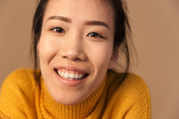 joyous asian woman wearing sweater smiling while posing at front in studio isolated over beige wall