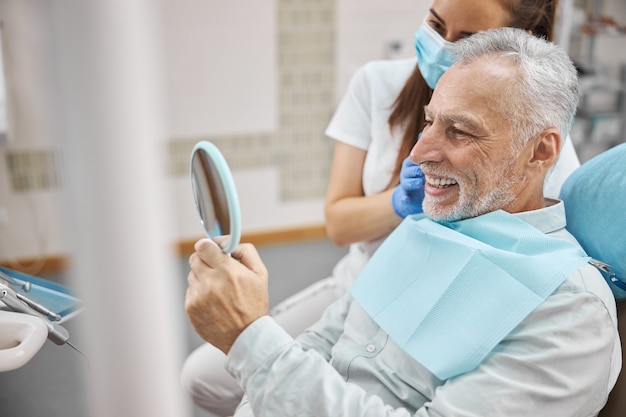 Photo joyous elderly man sitting in a dental chair with a doctor by his side while looking in the mirror