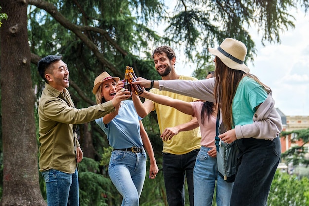 Joyous friends clinking beer bottles in a park celebrating together with laughter and happiness