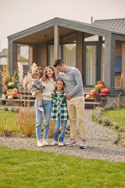 Photo joyous pretty teen girl posing for the camera with her little brother and parents against the house