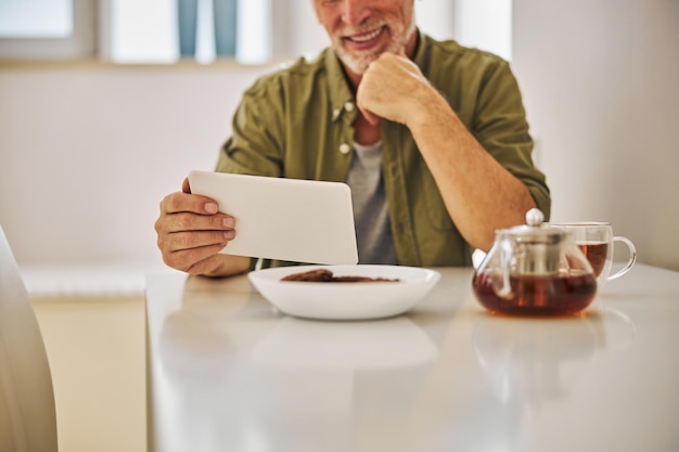 Anziano gioioso che beve tè mentre guarda il tablet