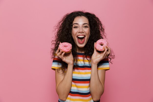 joyous woman 20s with curly hair having fun and holding donuts isolated on pink