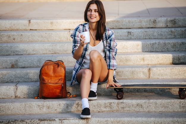 Joyous woman sitting on the stairs next to her skateboard having a rest
