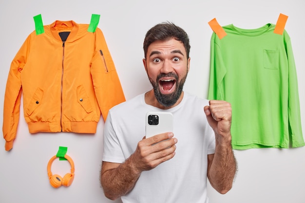 Jubilant happy bearded man clenches fist says yes uses mobile phone exclaims from joy wears white t shirt poses indoor with plastered jacket jumper and headphones Yes finally I gained success