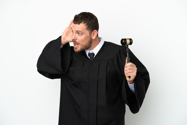 Judge caucasian man isolated on white background listening to something by putting hand on the ear