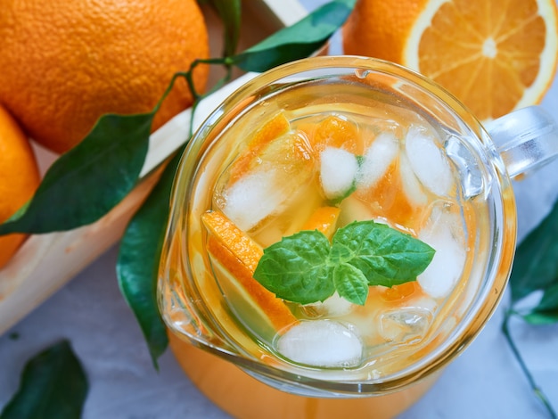 Jug of fresh orange juice with orange slices, ice, mint leaves on a gray surface