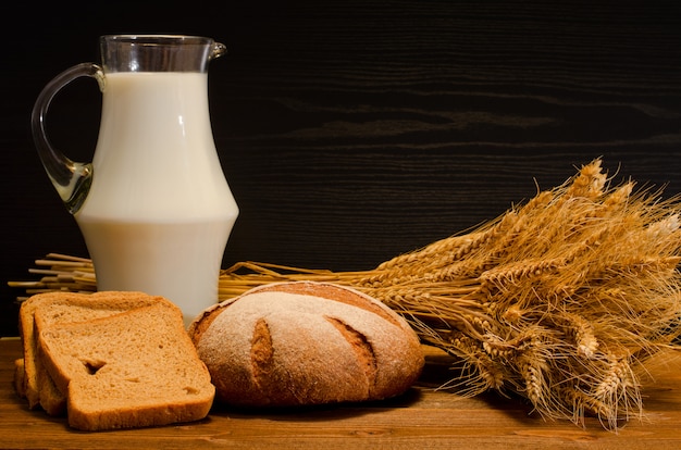 Jug of milk, round and square rye bread, a sheaf on wooden table, black background, space for text