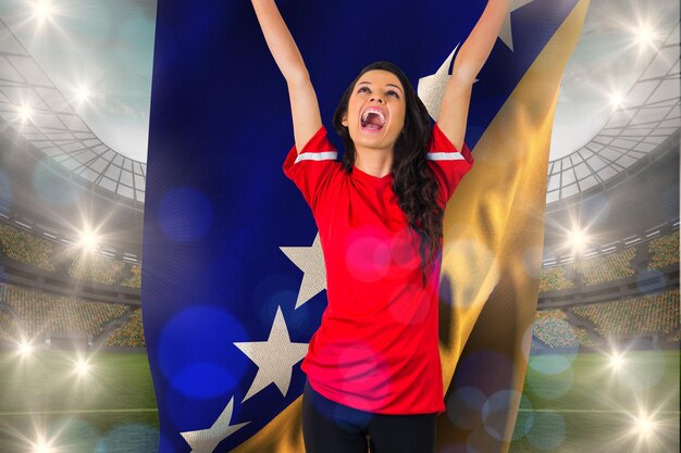 Juichende voetbalfan in het rood met de bosnische vlag tegen een groot voetbalstadion met lichten