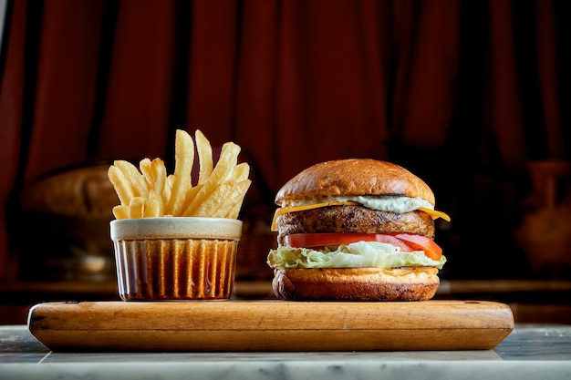 Juicy and appetizing burger with tomatoes, lettuce, cucumber, melted cheese and onions with brioche bun served on a wooden board with fries