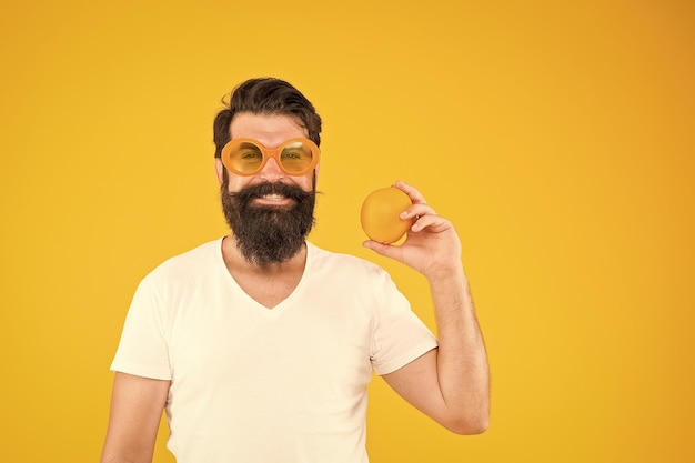 Juicy fruit Man bearded hipster in orange sunglasses on yellow background Cheerful guy with ripe fruit Summer vacation Fresh and healthy Summer nutrition Hipster with beard in summer mood