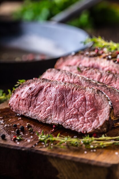 Juicy medium beef Rib Eye steak slices on wooden board with fork and knife herbs spices and salt