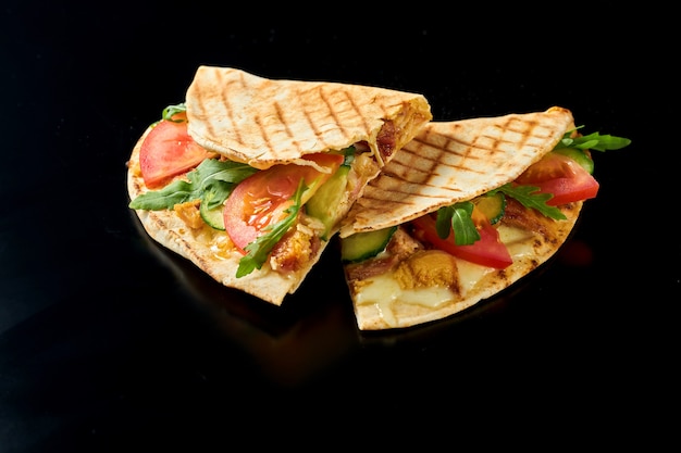 Juicy Mexican quesadilla with chicken, melted cheese, tomatoes, cucumber and arugula. Isolated on black surface. Street food. Close up, selective focus