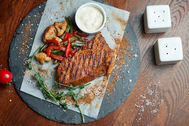 Juicy and mouth-watering grilled ribeye steak with spicy butter on a black tray with a side dish of vegetables. American cuisine.