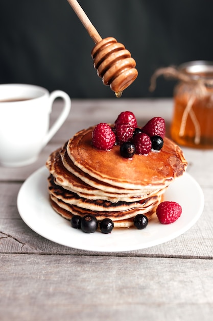 Juicy pancakes with berries and honey on a white plate, spoon, jar, wooden table, coffee cup.