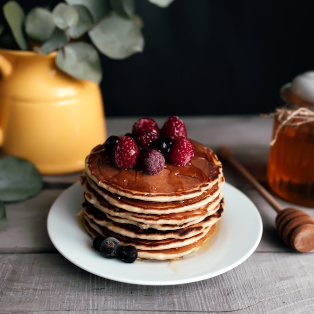 Juicy pancakes with berries and honey on a white plate, spoon, jar, wooden table, yellow vase with eucalyptus. High quality photo