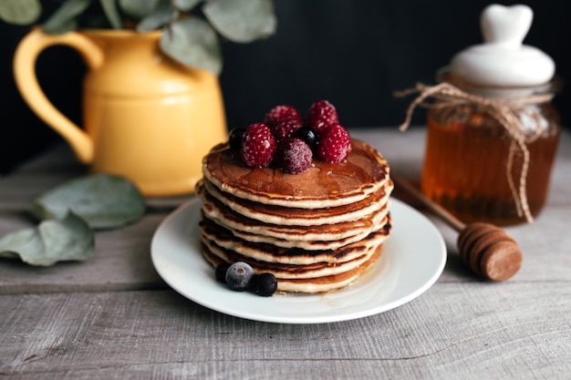 Juicy pancakes with berries and honey on a white plate, spoon, jar, wooden table, yellow vase with eucalyptus. High quality photo