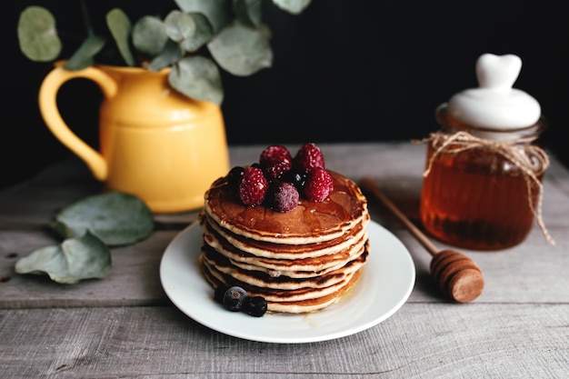 Juicy pancakes with berries and honey on a white plate, spoon, jar, wooden table, yellow vase with eucalyptus