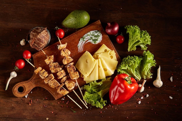 Juicy pork kebab on skewers laid out on a cutting board with fresh vegetables and cheese