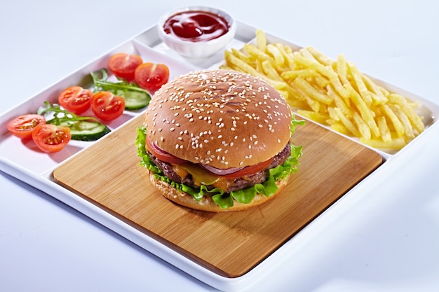 Juicy tasty hamburger on a wooden cutting board with french fried fries, vegetables and ketchup. Isolated composition on a white background and white serving tray.