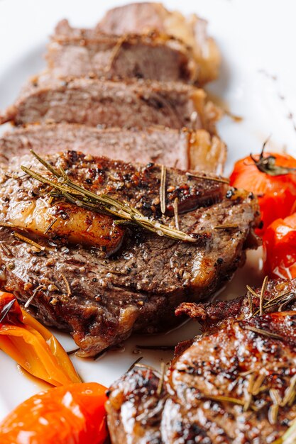 Juicy, well-done ribeye steaks on a white platter on a dark wooden table top with grammarine and fried cherry tomatoes and pepper