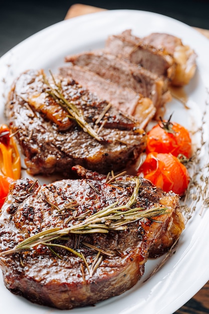 Juicy, well-done ribeye steaks on a white platter on a dark wooden table top with grammarine and fried cherry tomatoes and pepper