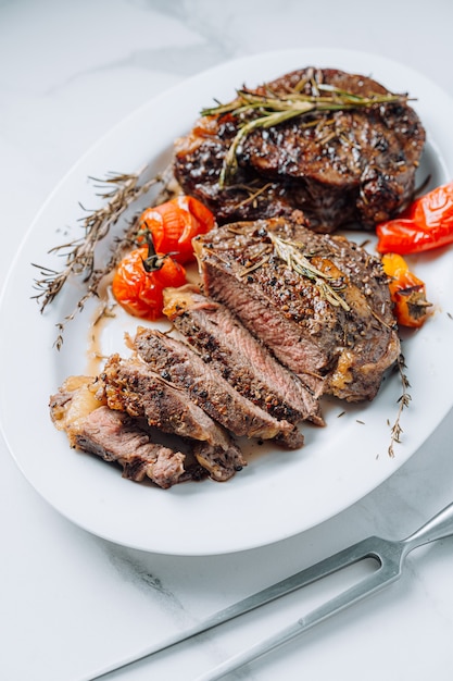 Juicy well-fried ribeye steaks on a white platter on a white marble surface with raspmarine and fried cherry tomatoes and peppers. steaks are sliced and ready to serve