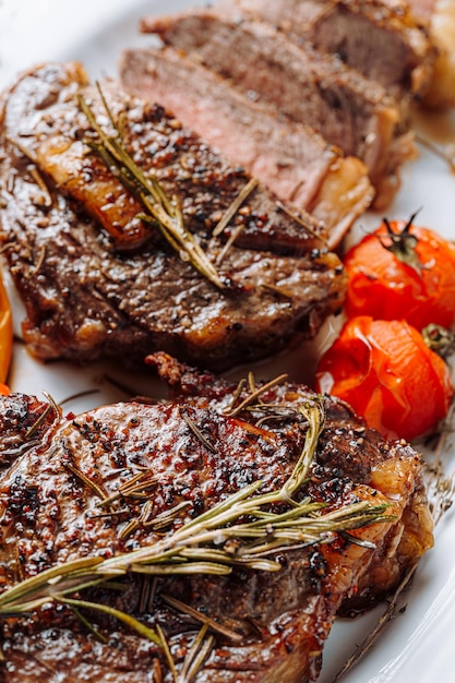 Succose bistecche di ribeye ben fatte su un piatto bianco su un tavolo di legno scuro con grammarina e pomodorini fritti e pepe