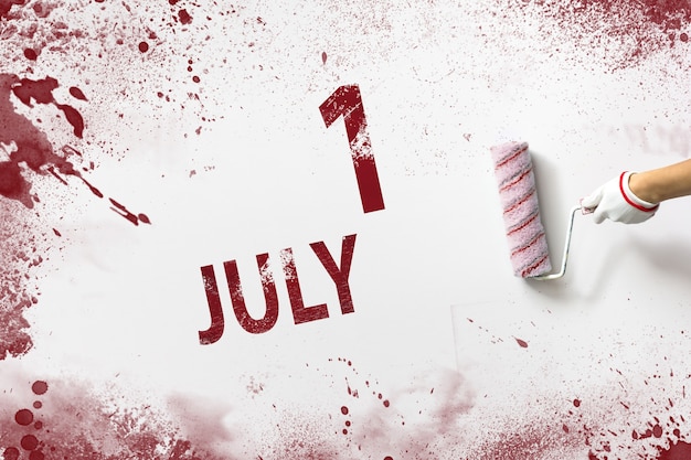 사진 7월 1일 매월 1일, 달력 날짜. 손은 빨간색 페인트로 된 롤러를 잡고 흰색 배경에 달력 날짜를 씁니다. 여름 달, 올해 개념의 날.