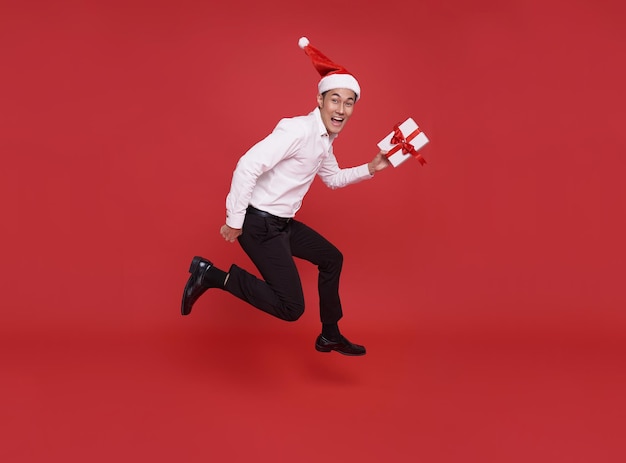 Uomo d'affari asiatico di salto che indossa il cappello di babbo natale che tiene una scatola regalo a natale e felice anno nuovo party