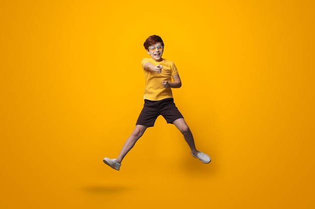 Jumping boy with red hair is wearing glasses and point at camera wearing a yellow shirt on a studio wall