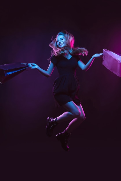 Jumping, flying high with shopping bags, laughting. Portrait of young woman in neon light on dark backgound. The human emotions, black friday, cyber monday, purchases, sales, finance concept.