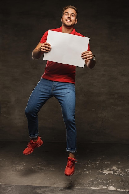 Jumping young man in jeans and red t shirt holding white sheet of paper.