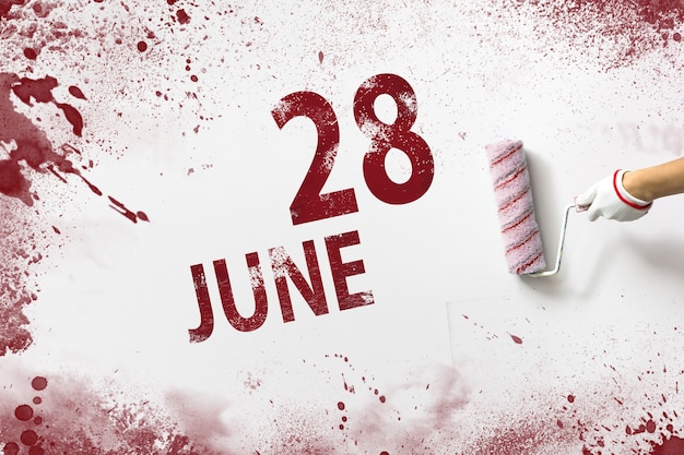 6月28日。月の28日、カレンダーの日付。手は赤いペンキでローラーを保持し、白い背景にカレンダーの日付を書き込みます。夏の月、年の日の概念。