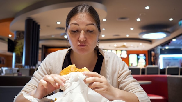 ジャンクフードのコンセプト。若いブルネットの女性は、フードコートのファーストフードレストランでハンバーガーを食べています。空腹の女の子がハンバーガーを噛んで噛んでいます。