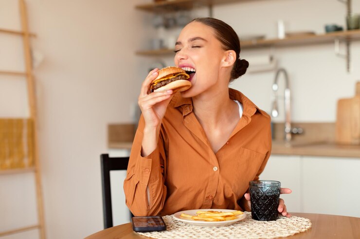 Photo junk food lady eating tasty burger holding and biting unhealthy fastfood sitting at table in kitchen