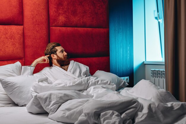 Foto appena sveglio, bell'uomo con lunghi baffi e barba, sdraiato a letto e guardando l'alba dalla finestra. concetto di mattina