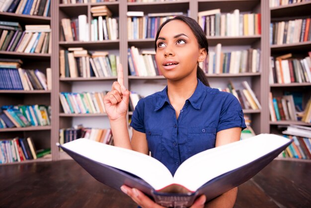 Just inspired. Surprised African female student holding a book and pointing up while sitting on the floor in library